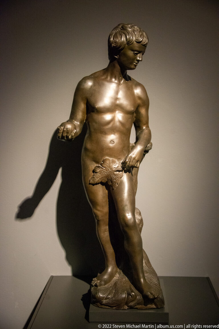 Small bronze nude male figure in contrapposto, lit dramatically so that a large shadow falls on the wall behind.