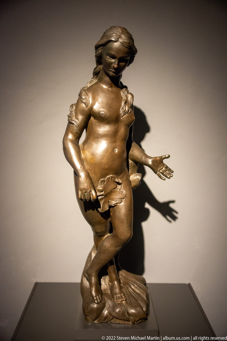 Small bronze nude male figure in a twisting stance on a modeled base, lit strongly from the side.