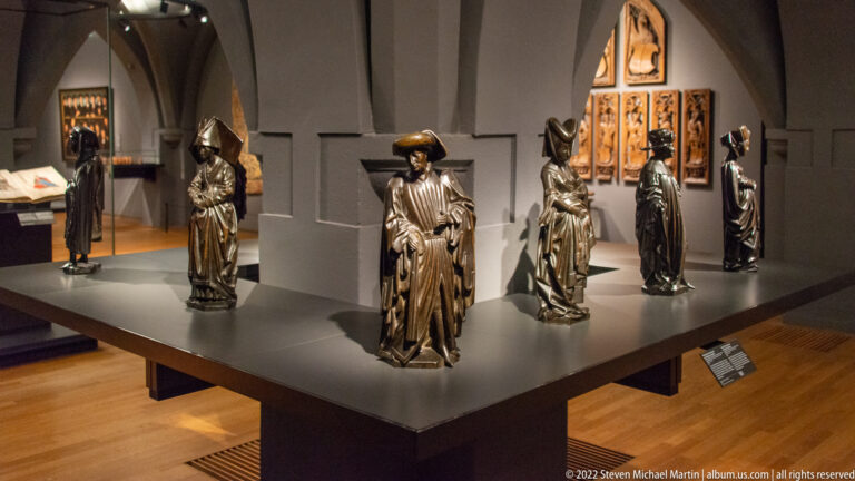 Six small metal statuettes in historical dress arranged on a dark museum platform before a carved panel.