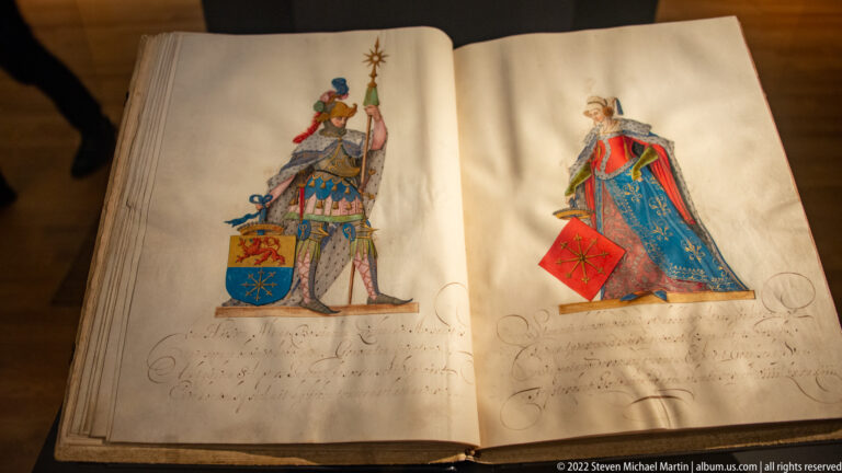 Open manuscript with two colorful heraldic figures, coats of arms, and handwritten annotations.