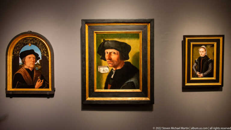 Three framed historical portraits of men and a woman arranged on a dark museum wall.