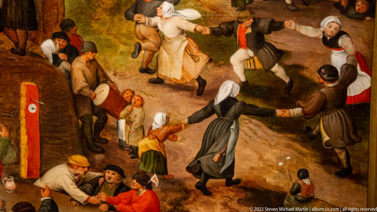 Villagers in historical dress hold hands and dance energetically outdoors while other figures watch nearby.