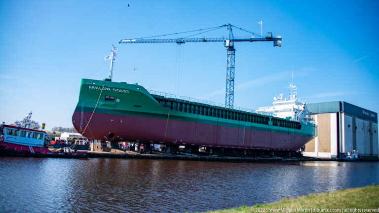 Large green-and-red cargo ship out of the water beside a crane, shed, and smaller service vessels.