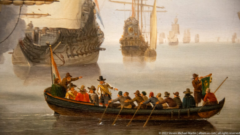 Long oared boat carrying formally dressed passengers between large anchored sailing ships.