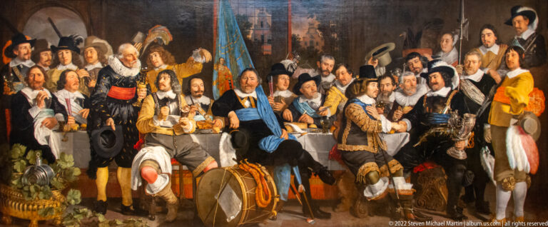 Large militia company in seventeenth-century dress gathered around a banquet table with banners, drums, and weapons.