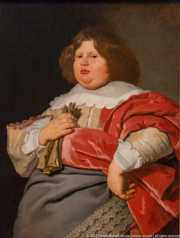 Seated young sitter in a red satin cloak and pale lace-trimmed clothing against a dark background.