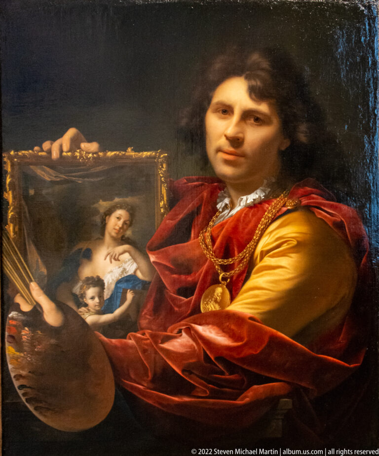 Male painter in red and gold clothing holding a palette and a small framed painting of a woman and child.