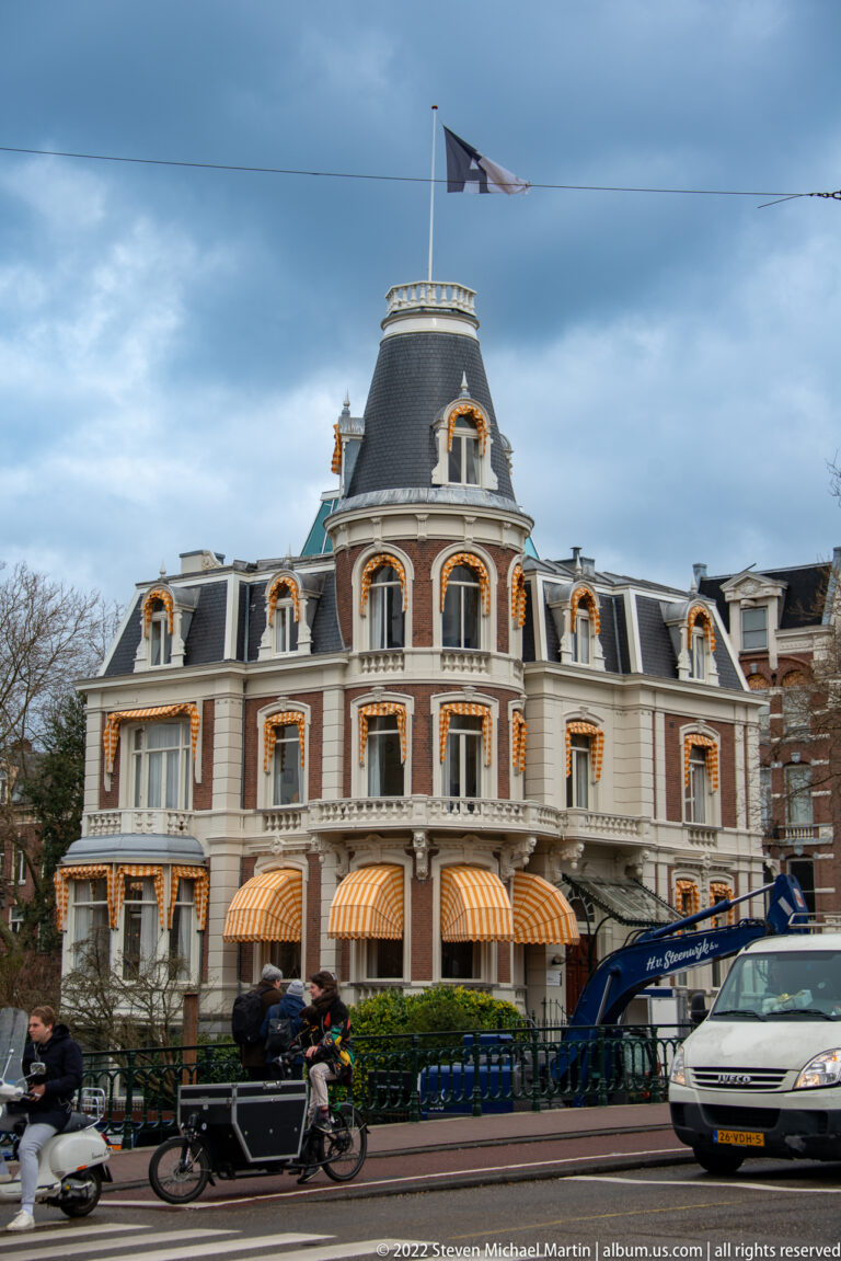 Grand pale corner building with mansard roof, ornate trim, striped awnings, flag, bicycles, and street traffic.