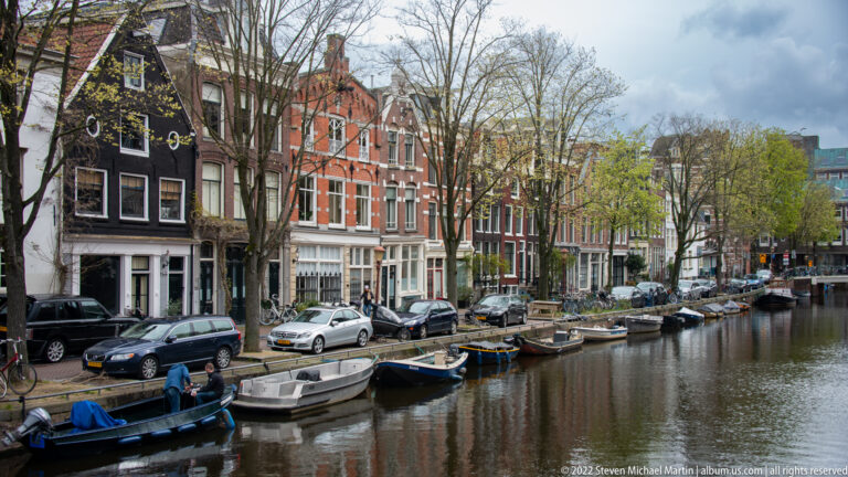 Tree-lined Amsterdam canal bordered by narrow historic houses, moored small boats, parked cars, and bicycles.
