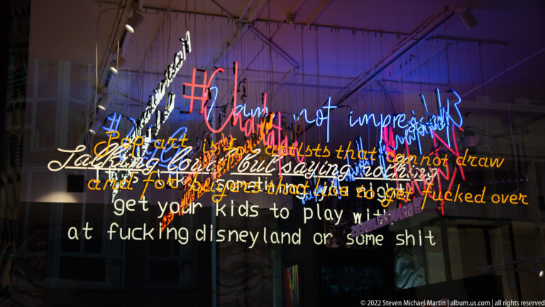 Layered multicolored neon text glows across a dark glass surface, with overlapping handwritten phrases and reflections.