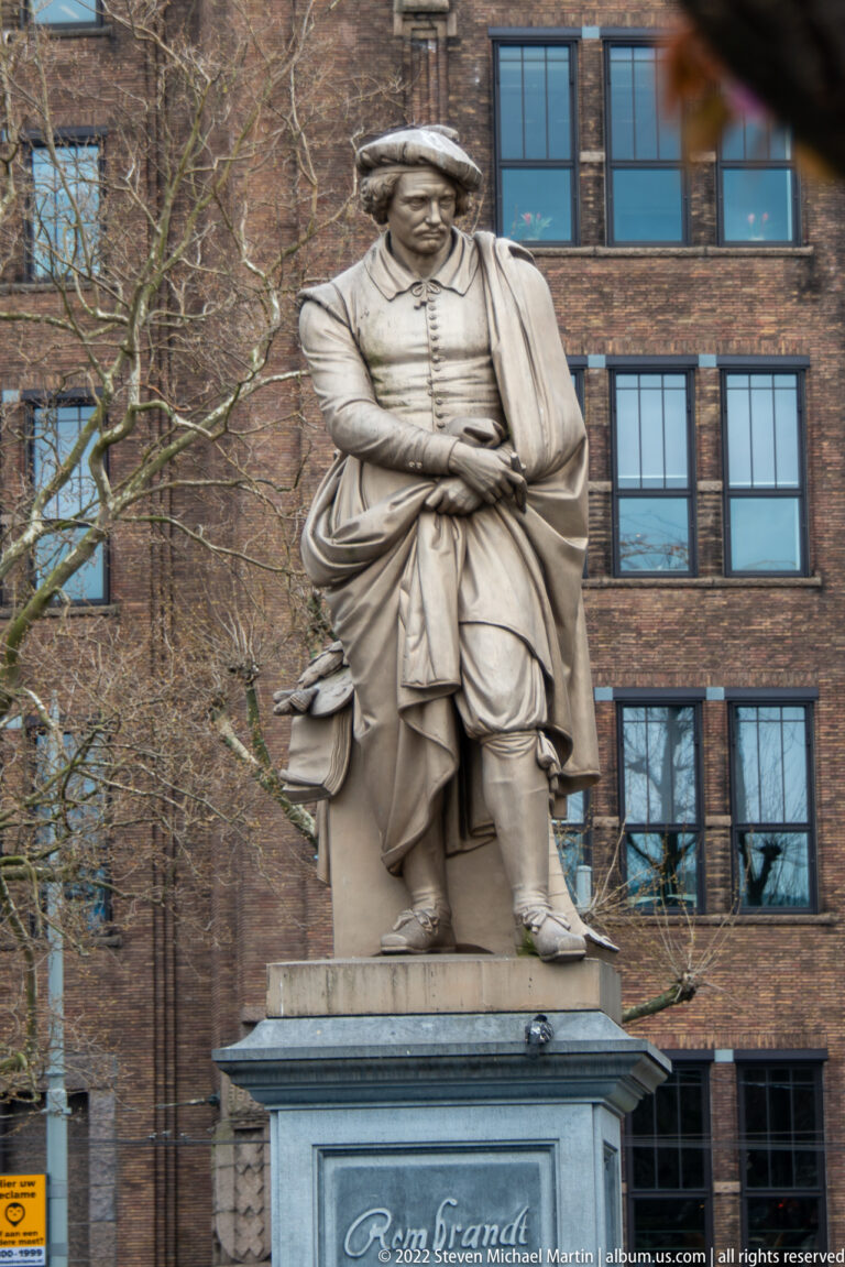 Stone statue of Rembrandt in historical dress on a pedestal before a brick building and leafless trees.