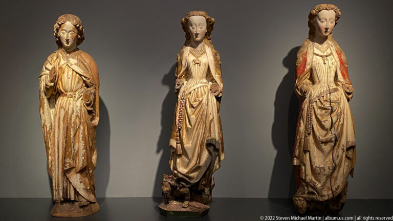 Three standing polychrome female religious sculptures in long gilded gowns displayed against a dark wall.