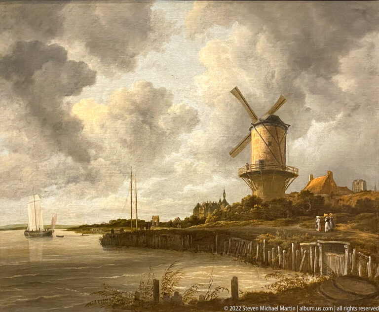 Dutch landscape painting with a large windmill beside water, sailboats, low shoreline, and cloudy sky.
