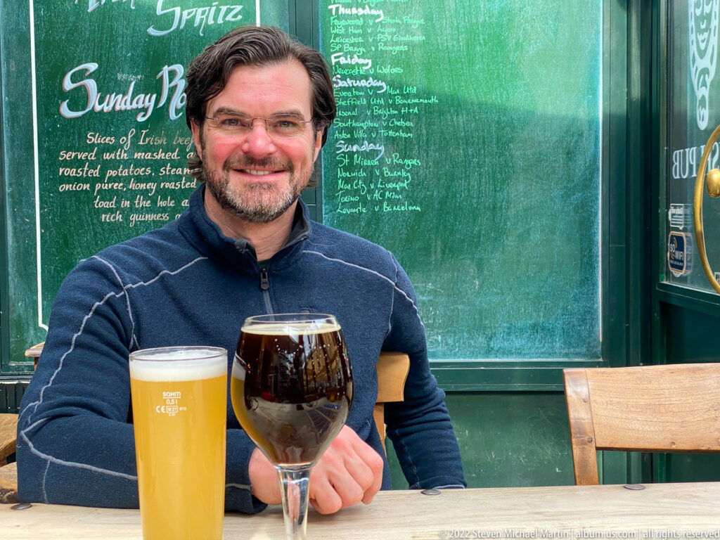 Smiling man at an outdoor café table with one pale beer and one dark beer before a chalkboard menu.