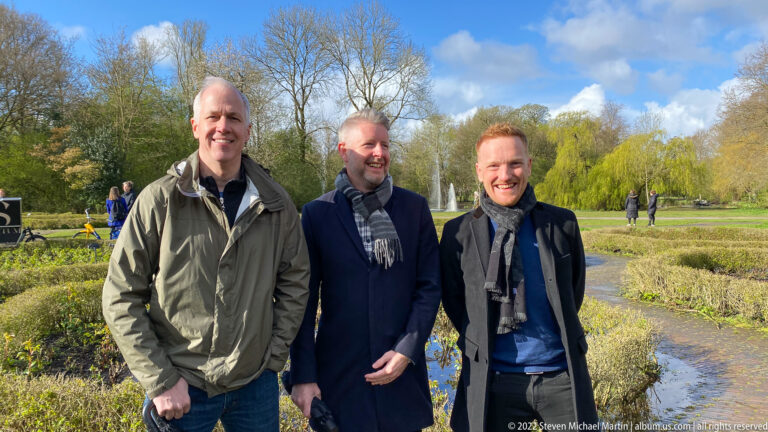 Three smiling men standing together beside water and planted beds in a leafy Amsterdam park.