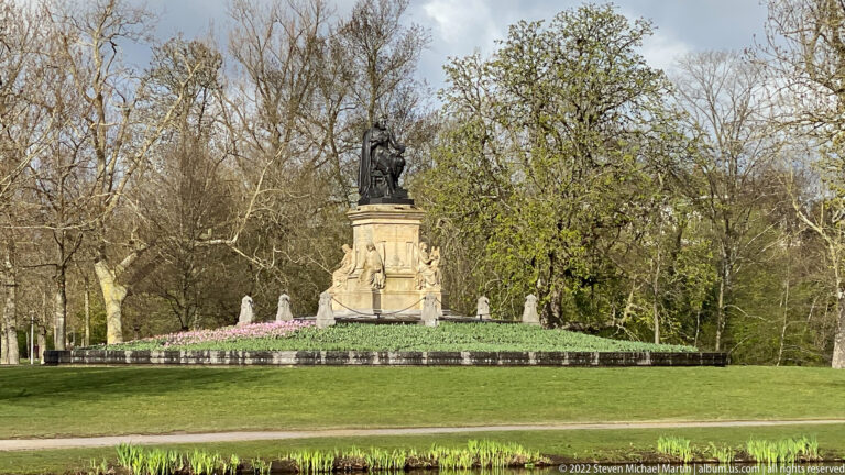 Bronze figure monument on a tall stone pedestal across a lawn, framed by trees and spring flower beds.