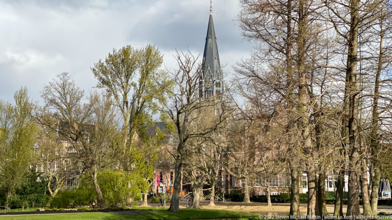 Slender Gothic Revival church spire rising behind leafless and budding trees in an Amsterdam park district.