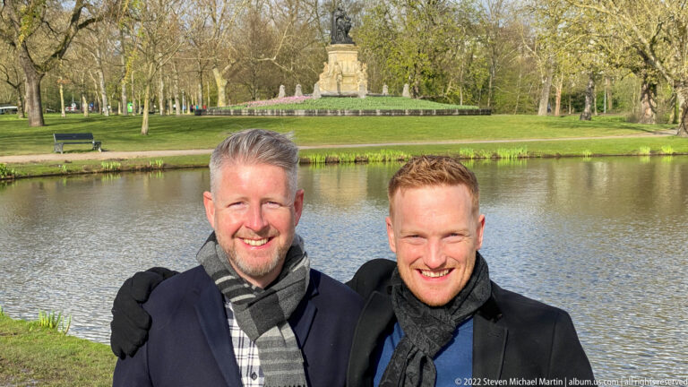 Two smiling men beside a park pond, with lawns, trees, flower beds, and a monument in the distance.