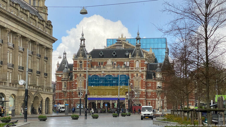 Ornate red-brick theater with steep roofs, turrets, pale trim, and banners at the end of an Amsterdam square.