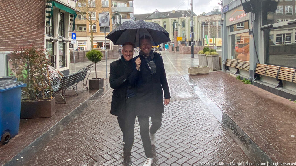 Two men walking under a black umbrella along a wet brick street lined with shops, benches, and planters.