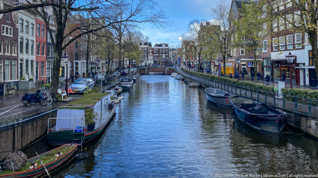 Amsterdam canal lined with moored boats, bicycles, trees, storefronts, and narrow historic buildings.