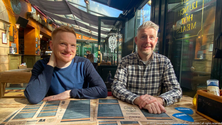 Two smiling men seated at a café table with menus, warm woodwork, glass, and pub signage around them.