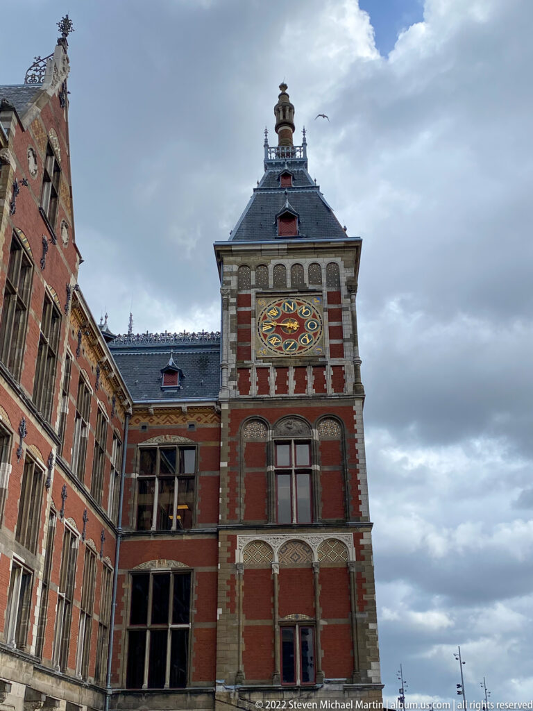 Ornate Amsterdam Centraal tower with red brick, pale stone trim, steep roof, and a large colorful circular dial.