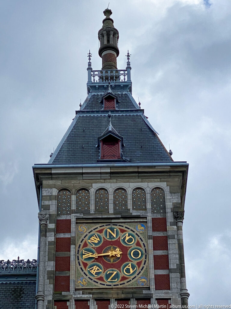 Close view of Amsterdam Centraal's ornate circular dial beneath a steep roof and small rooftop cupola.