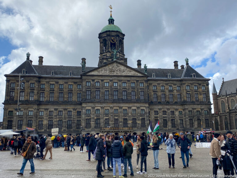 Royal Palace Amsterdam across Dam Square, with symmetrical stone façade, central cupola, clock, and crowds in front.