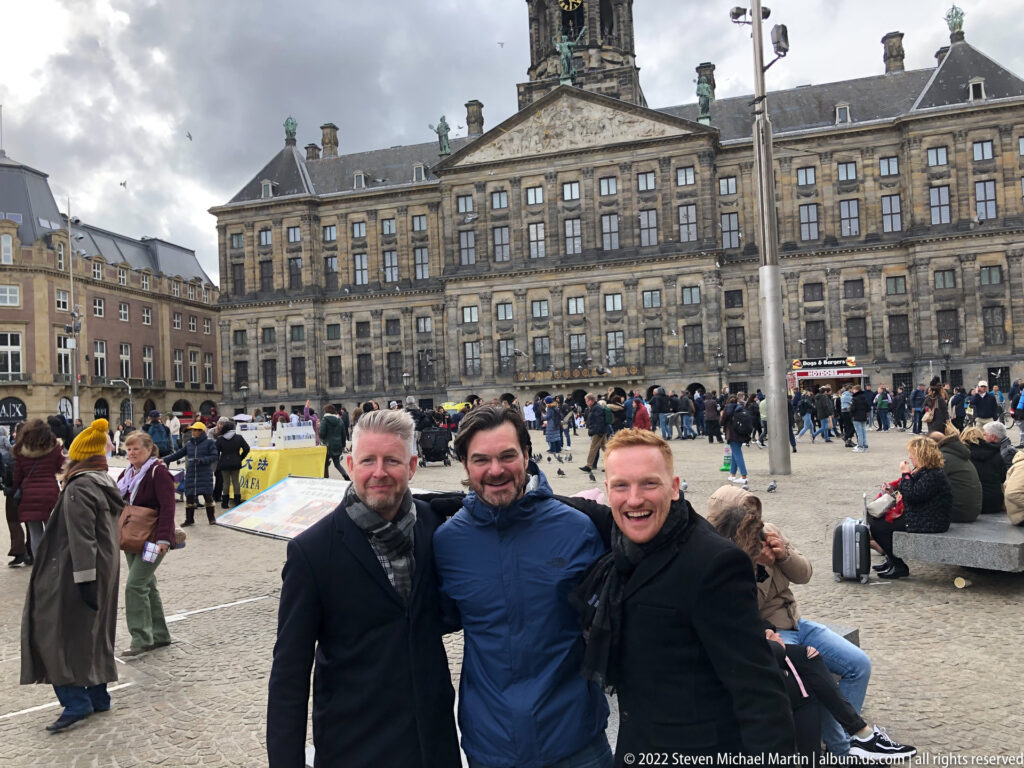 Three men posing on crowded Dam Square with the Royal Palace Amsterdam spanning the background.