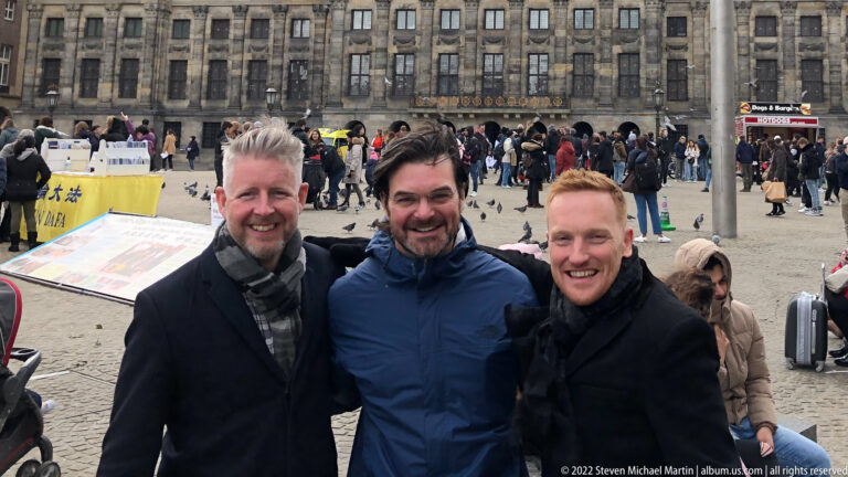 Close group portrait of three men on Dam Square with crowds and the Royal Palace Amsterdam behind them.