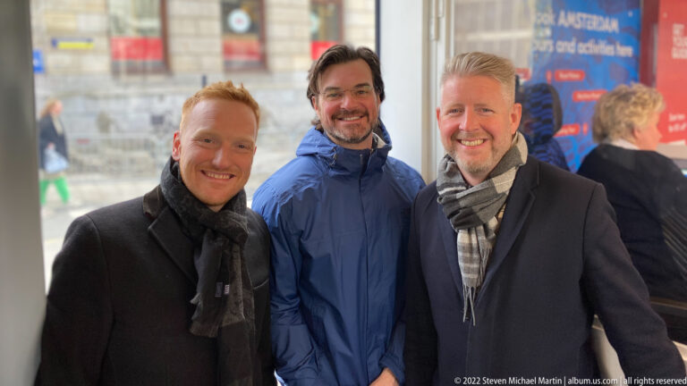 Three men posing together inside a bright modern interior with blurred transit-style signage and glass behind them.
