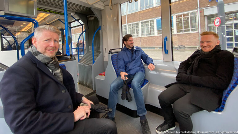 Three men seated inside a modern tram with blue grab rails, gray seats, large windows, and articulated joint.