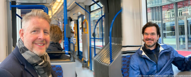 Two smiling men seated inside a modern tram with blue grab rails, patterned seats, windows, and an articulated joint.
