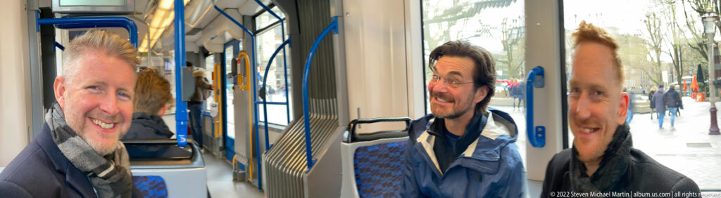 Three men gathered inside a tram near the doorway, with blue rails, patterned seats, and pedestrians visible outside.
