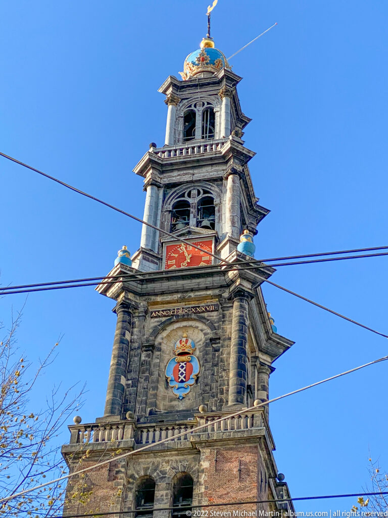 Upward view of the Westerkerk's Westertoren with clock faces, heraldic ornament, gilded crown, and tram wires.