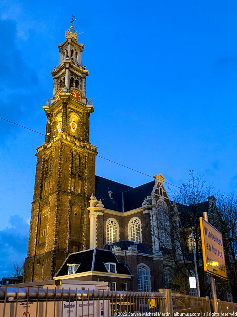 Westerkerk and illuminated Westertoren at dusk, with glowing clock face, church nave, barriers, and blue evening sky.