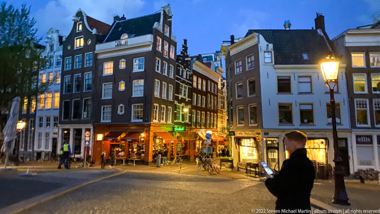 Blue-hour Amsterdam street corner with narrow buildings, cafés, bicycles, glowing windows, streetlamp, and a man using his phone.