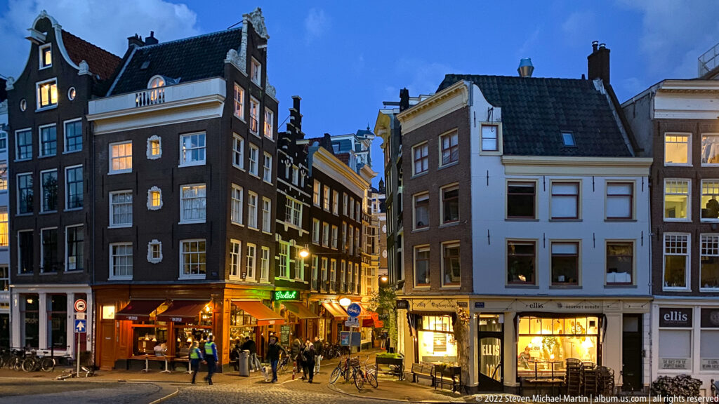 Historic Amsterdam street corner at blue hour with narrow gabled buildings, restaurants, pedestrians, and bicycles.