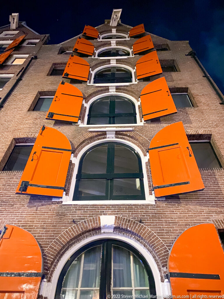 Upward view of a narrow brick Amsterdam façade with stacked arched windows and vivid orange shutters.