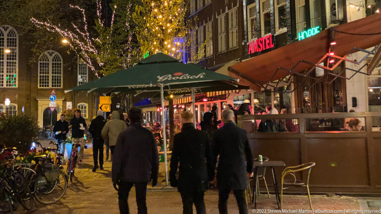 Busy Amsterdam night street with café terrace, pedestrians, bicycles, decorative tree lights, and illuminated beer signs.