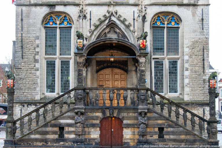 Close view of Gouda Town Hall entrance with twin stone stairs, Gothic porch, heraldic shields, statues, and tall windows.
