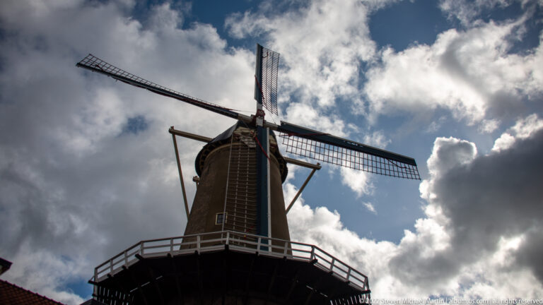 Low-angle view of De Roode Leeuw windmill with four sails and gallery against a cloudy sky.