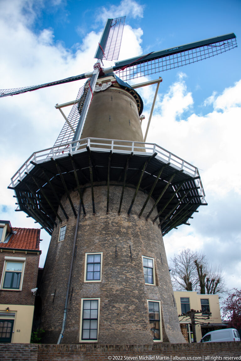 Brick tower of De Roode Leeuw windmill with timber gallery, four sails, and neighboring rooftops.