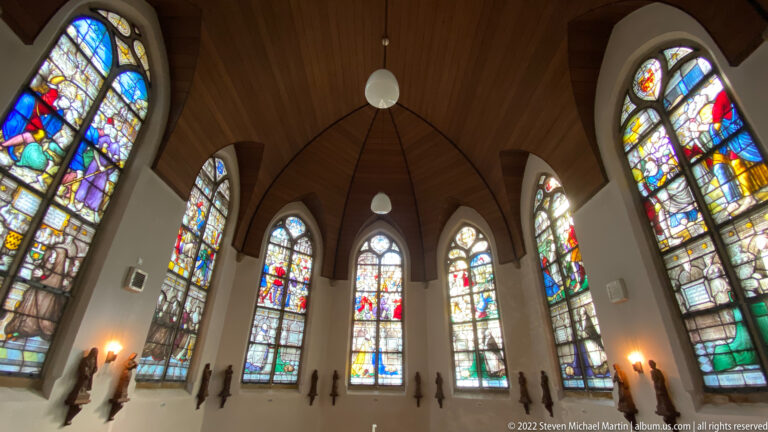Curved chapel interior with tall stained-glass windows, timber ceiling, pendant lights, and dark wall memorials.