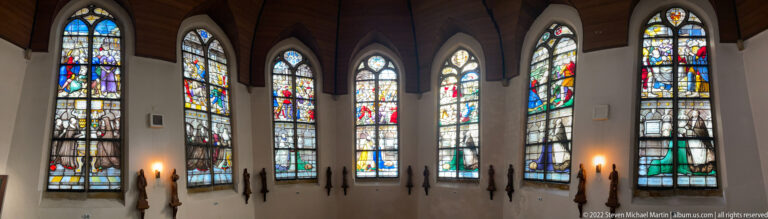 Six tall stained-glass windows arranged around a curved chapel wall with dark memorial forms below.