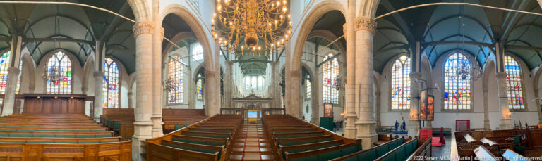 Central nave of Sint-Janskerk with wooden benches, pale columns, green vaulting, chandeliers, and stained glass.