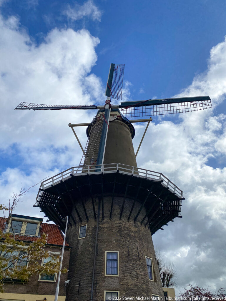 Low-angle view of De Roode Leeuw windmill with brick tower, broad gallery, and sails against blue sky.