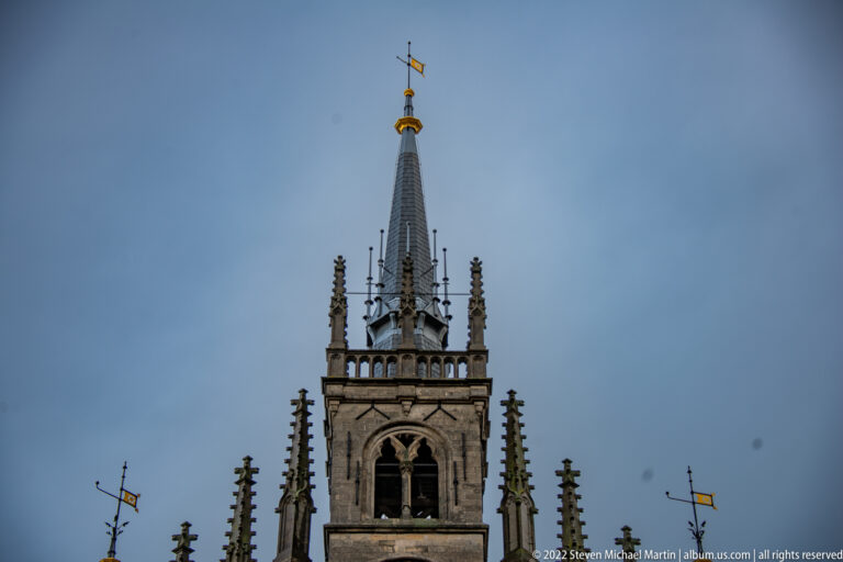 Upper tower and spire of Gouda Town Hall with belfry openings, pinnacles, gilded finial, and weather vane.