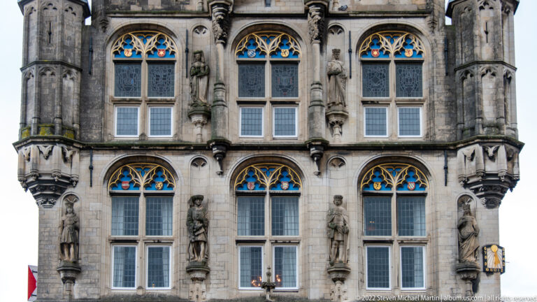 Detail of Gouda Town Hall façade with tall traceried windows, carved standing figures, blue-and-gold accents, and stone ornament.
